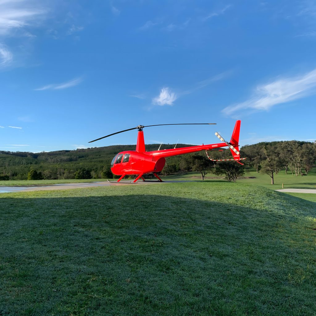 Bright red R44 helicopter sitting on a concrete helipad, with green grass and trees in the background, and blue sky.