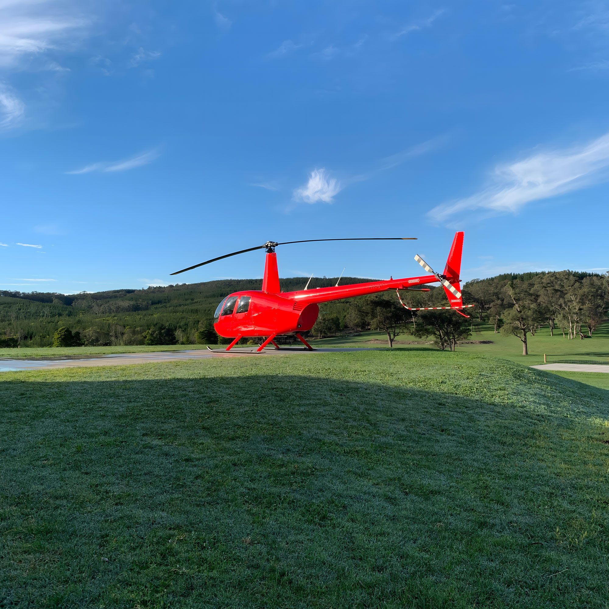 Bright red R44 helicopter sitting on a concrete helipad, with green grass and trees in the background, and blue sky.
